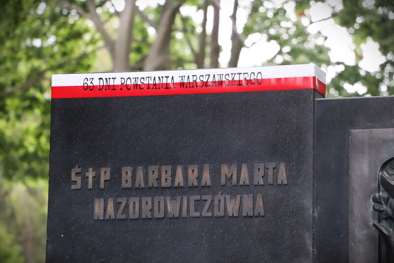 Fotografia przedstawia fragment grobu Barbary Nazdrowicz uczestniczki Powstania Warszawskiego. W górnej części tablicy nagrobnej umieszczona jest biało-czerwona szarfa z napisem sześćdziesiąt dni Powstania Warszawskiego.