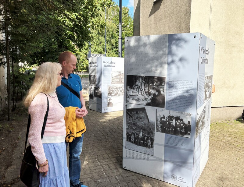Fotografia przedstawia osoby odwiedzające wystawę poświęconą życiu i działalności św. o. Maksymiliana Marii Kolbego. Wystawa znajduje się przed budynkiem kościoła pw. Matki Bożej Anielskiej w Łodzi. Wystawa składa się z kilku modułów, na których zawarte są fotografie oraz tekst dotyczący Św. Maksymiliana.